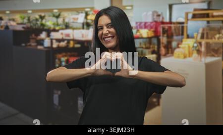 Donna che crea una forma a cuore con le mani e sorride tra esposizioni di profumi in un negozio di vendita al dettaglio di edifici; gioiosa celebrazione dell'affetto. Foto Stock