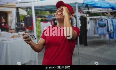 Uomo che tiene lo smartphone con entrambe le mani nelle bancarelle del mercato di strada, indossa un berretto rosso e una maglietta rossa, ridendo e sorridendo; divertimento di gioco. Foto Stock