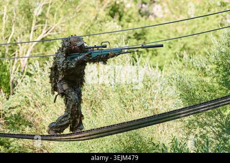 Cecchino soldato in Ghillie Suit Going Rifle mentre attraversa il ponte sospeso di corda durante l'addestramento militare di ricostruzione Foto Stock