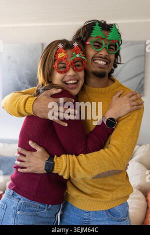 Coppia che abbraccia e ride in salotto indossando occhiali festivi, maglioni in maglia e smartwatch. Abbracciare, coppia, casa, accogliente, holi Foto Stock