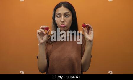 Donna che tiene due fragole tra le dita con mani visibili sullo sfondo arancione dello studio; indecisione. Foto Stock