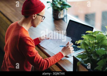 Freelance uomo in occhiali rosso maglione lavoro a distanza con il computer portatile seduto in un bar che lavora con documenti Foto Stock