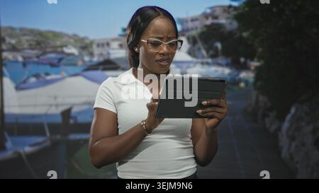 Donna che tiene in mano un tablet e sta girovagando mentre legge con le dita sullo schermo in uno studio con sfondo porticciolo e oggetti di scena del molo; attenzione all'esame. Foto Stock