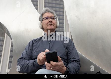 Uomo asiatico anziano seduto su una scultura di metallo in plaza, che usa lo smartphone con le cuffie bianche. Urbano, paesaggio urbano, opere d'arte, riflessi Foto Stock