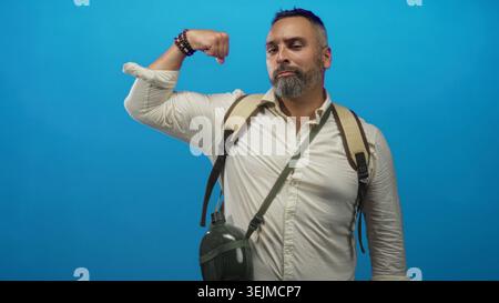 L'uomo con zaino e mensa flette l'avambraccio e i gesti con la mano in blu studio, ispanico di mezza età con barba grigia; sicurezza all'avventura. Foto Stock