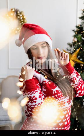Giovane donna con cappello e maglione di babbo natale che regge splendide luci di natale Foto Stock
