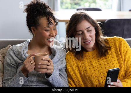 Diverse amiche che siedono sul divano nel soggiorno, tenendo in mano smartphone e tazza in ceramica. Donne, amicizia, accogliente, casa, telefono, Foto Stock