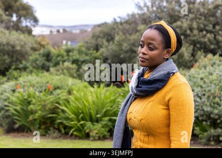 Donna afroamericana in piedi nel parco con maglione giallo senape e sciarpa grigia, copia spazio. Giardino, esterno, verde Foto Stock