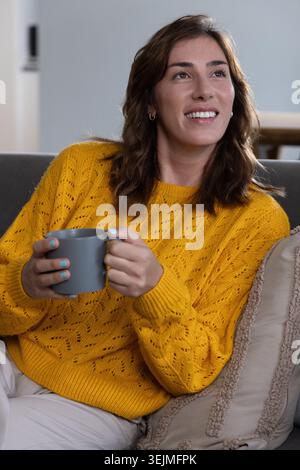 Donna seduta sul divano nel soggiorno, indossa un maglione giallo senape e tiene in mano una tazza grigia opaca. Accogliente, casa, luce del giorno, rilassato, Foto Stock