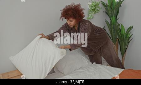La donna mette il cuscino sul letto e leviga le lenzuola bianche mentre nasconde un tiro arancione accanto a una testata di legno in una camera da letto dell'edificio; calma routine. Foto Stock