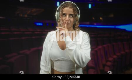 Donna con cuffie, dita verso le labbra per il silenzio in un auditorium con cappuccio bianco e ritaglio superiore; messa a fuoco tranquilla. Foto Stock