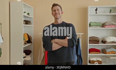 Uomo con le braccia incrociate che indossa un maglione in maglia in un guardaroba da studio, in piedi accanto a un scaffale e scaffale; sicuro di sé. Foto Stock