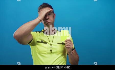 L'arbitro uomo tiene in mano il cartellino giallo, mano a fronte, indossa fischietto in blu studio; frustrazione di rigore di gioco. Foto Stock