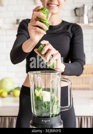 Giovane donna bionda sorridente che prepara frullati di cetrioli nella cucina di casa Foto Stock