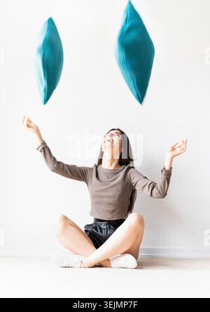 Bella donna felice in abiti informali seduti sul pavimento che getta cuscini in aria Foto Stock