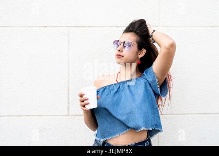 Giovane donna elegante che indossa camicia jeans, occhiali da sole e borsa, beve caffè per andare in strada, sfondo bianco sulle pareti Foto Stock