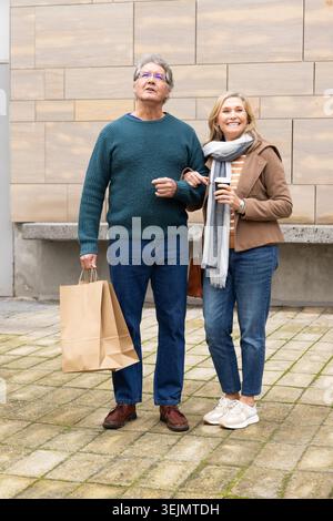 La coppia senior si trova sulla piazza lastricata di fronte alla facciata beige, tenendo in mano una borsa della spesa e un caffè. Anziani, compagnia, città, Foto Stock