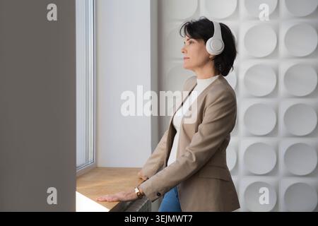 Donna di mezza età che indossa le cuffie rilassata e che fa meditazione seduta su una scrivania in ufficio. concetto senza stress. mentale Foto Stock