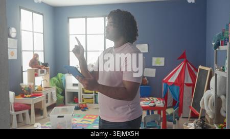 Uomo con tablet tocca lo schermo e punta il dito nell'edificio della sala giochi dell'asilo; calma apprendimento della concentrazione. Foto Stock