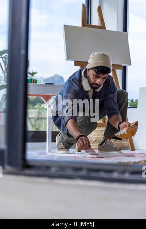Artista maschile accovacciato su tela da pavimento con tavolozza, indossando berretto e cuffie. Cavalletto, pennello, pittura, atelier, Foto Stock