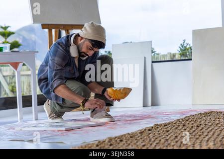 Uomo barbuto accovacciato su un foglio in studio, indossando un berretto, applicando vernice dalla ciotola alla tela del pavimento. Artista, minimalista, luce del giorno Foto Stock