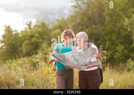 Due donne di mezza età sono amiche con zaini che guardano la mappa in piedi sulla montagna, camminano nella natura e trascorrono del tempo Foto Stock
