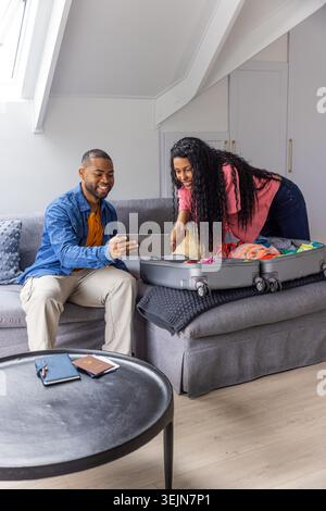 Coppia che prepara la valigia aperta con ruote su un divano grigio nel loft, uomo che tiene un tablet, passaporto visibile. Valigia, viaggio, trasporto, piano Foto Stock