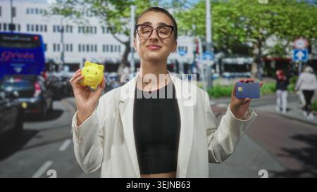 Donna dai capelli corti che tiene in mano un salvadanaio giallo e una carta di credito in una strada trafficata in città con giacca bianca e top nero; sicura di sé. Foto Stock