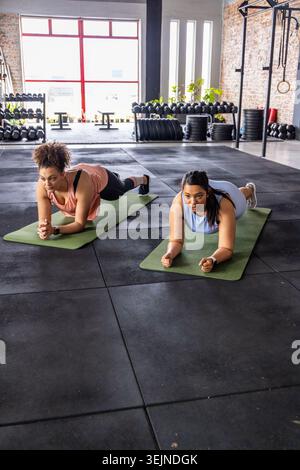Due donne che eseguono tavole dell'avambraccio su tappetini verdi in palestra indossando abiti sportivi con cremagliere. Studio, pesi, fitness, coppia, io Foto Stock