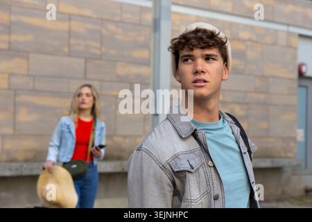 Amici adolescenti in piedi per strada urbana con giacche in denim leggero, con smartphone e cappello di paglia. Giovani, casual, zaino, esterno Foto Stock