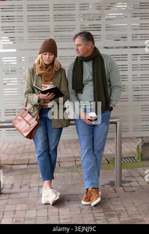 Coppia di mezza età in piedi presso una fermata di transito coperta, donna che scrive su un taccuino, uomo che tiene il caffè. Duo, diario, coppa, urban, st Foto Stock