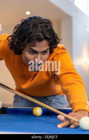 Uomo indiano degli anni Venti che indossa un maglione arancione appoggiato sul tavolo da biliardo in feltro blu a casa, puntando il segno. Biliardo, concentrazione, AM Foto Stock