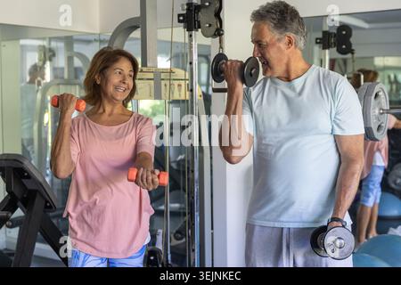Accoppiare il sollevamento manubri e sorridere mentre ci si allena in palestra con specchi e portapacchi. Fitness, allenamento, duo, senior, matur Foto Stock