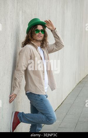 Uomo elegante con cappello verde di festa appoggiato al muro Foto Stock