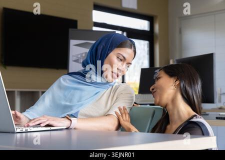 Diverse collaboratrici che collaborano alla scrivania in ufficio, una che indossa hijab, utilizza laptop e monitor. Collaborazione, lavoro di squadra, mese Foto Stock