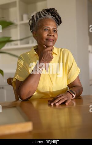 Donna anziana seduta al tavolo da pranzo in legno nella cucina di casa, luce solare su camicetta gialla, smartwatch. Caldo, contemplativo, sereno, Foto Stock