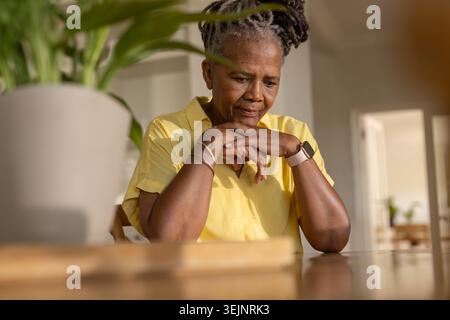 Donna afroamericana anziana seduta a un tavolo di casa, con la parte superiore gialla accanto alla pianta, con il mento a riposo. Contemplativo, serenità, por Foto Stock