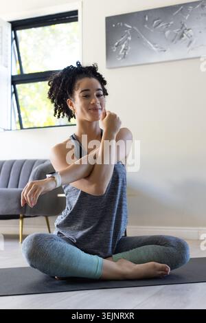 Donna adulta che si allunga il braccio lungo il petto sul tappetino da yoga a casa, grazie allo smartwatch. Fitness, benessere, stile di vita, Foto Stock