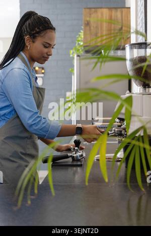 Barista afroamericana che prepara l'espresso dietro il bancone, indossa grembiule e smartwatch. Caffè, caffè, caffè, caffè, caffè, portafiltro Foto Stock