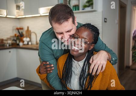 Felice coppia interrazziale che si abbraccia nella cucina di casa Foto Stock