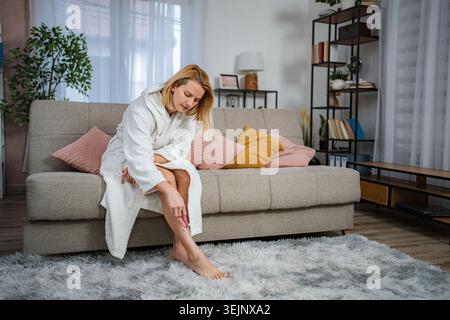 Giovane donna che rasa la gamba sul divano in accappatoio Foto Stock