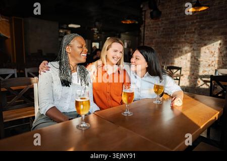 Tre diverse amiche donne che si godono la birra e ridono in un pub Foto Stock
