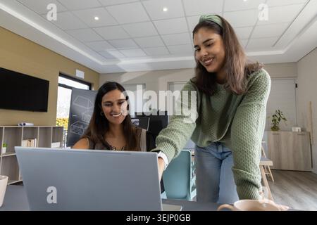 Diverse collaboratrici che si appoggiano al laptop con un caffè in ufficio, puntando sullo schermo. Collaborazione, donne, sala riunioni, centro benessere Foto Stock