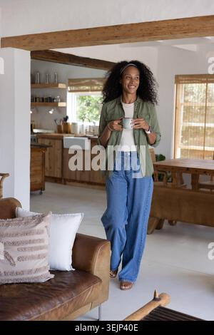 La persona tiene in mano una tazza di ceramica bianca in una zona soggiorno-cucina a pianta aperta, con travi di legno esposte. Rustico, accogliente, casalingo, natura Foto Stock