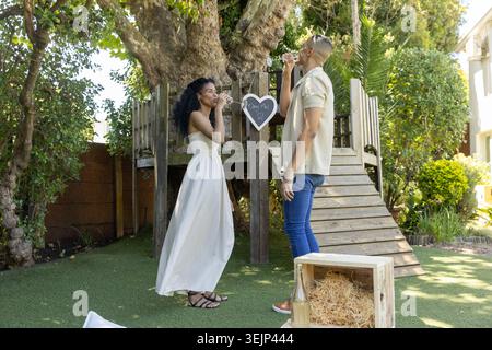 Coppia in piedi sorseggiando del vino nel cortile accanto alla casa sull'albero con il segno del cuore, donna in abito bianco. Romantico, all'aperto, giardino, illuminato dal sole, Foto Stock