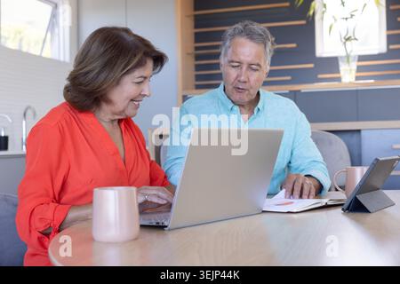 Donna anziana in camicetta di corallo che scrive su un notebook argento mentre l'uomo controlla il notebook al tavolo della cucina. Collaborazione, pianificazione, casa Foto Stock