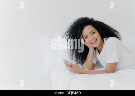 Felicissima, rilassata, riccia, giovane afroamericana gode del tempo libero a casa, si trova a letto con un moderno cellulare, usa il telefono Foto Stock