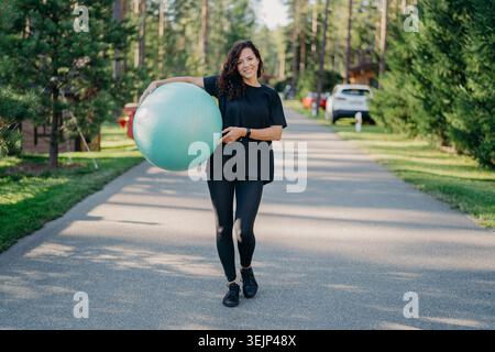 Giovane donna bruna in forma tiene una grande palla fitness, vestita con abbigliamento attivo, fa esercizio fisico, posa nel parco, passeggiate all'aperto e Foto Stock