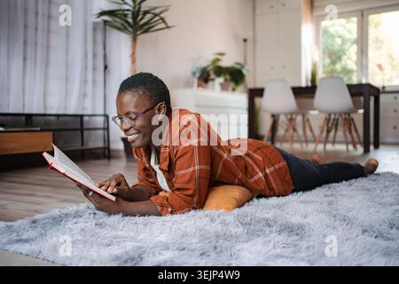 Giovane donna sdraiata sul tappeto che legge un libro a casa Foto Stock
