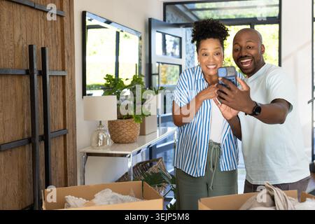 Coppia afroamericana che sorride e tiene in mano lo smartphone mentre disimballa le scatole in movimento nell'atrio di casa Foto Stock